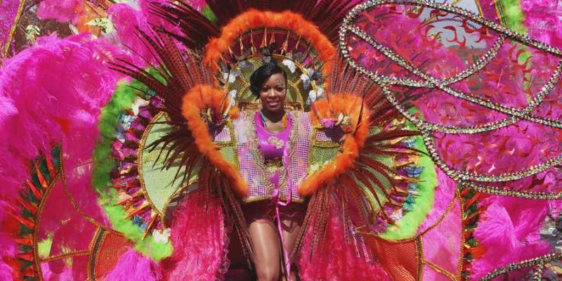 Participant in vibrant carnival costume featuring large feathered wings in pink, orange, and yellow, smiling at the camera.