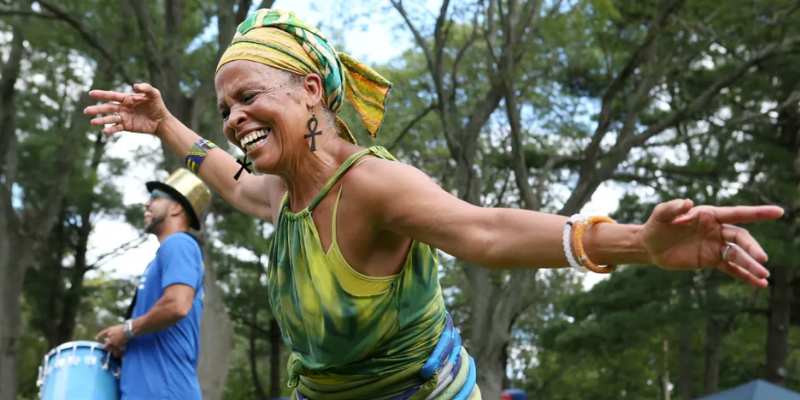 A joyful person dances to drum music at an outdoor event, wearing a green and yellow top, a colorful headscarf, and bracelets.