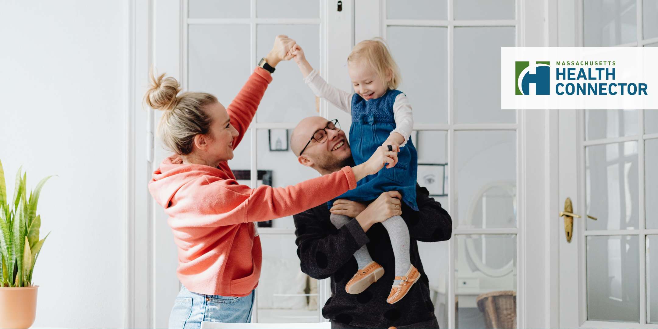 A joyous family in a living room with a father carrying his daughter on his shoulder as his wife reaches for the little girl as if dancing together.