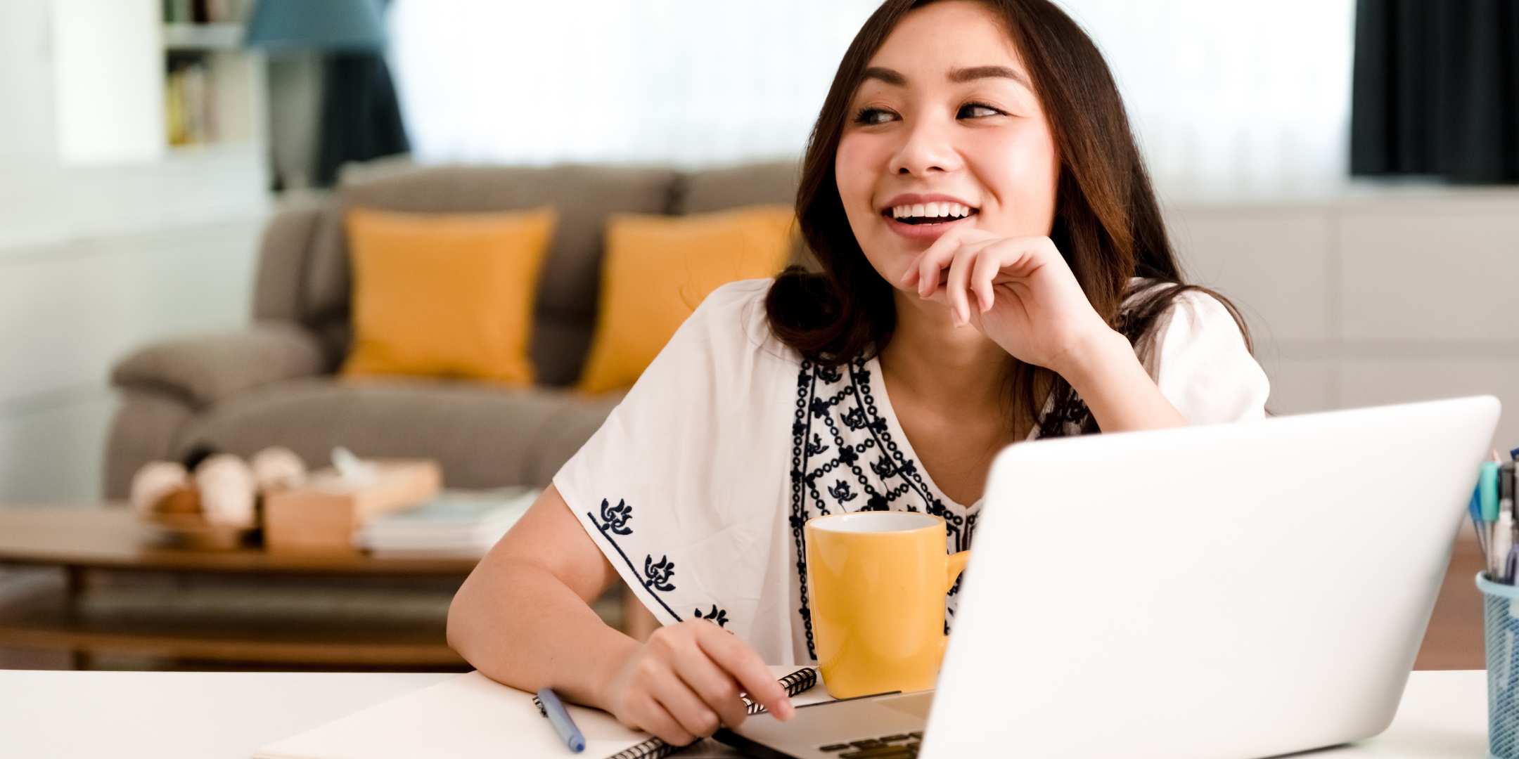 A person sitting at a table with a laptop, holding a yellow mug, smiling and looking away from the screen in a bright, cozy room.