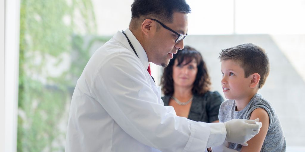 A young boy sitting on an examining table getting a shot in his arm by a doctor in an office with his mother.